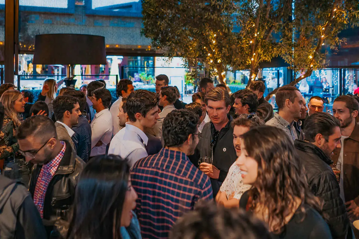 A cheerful crowd of real estate professionals talking and smiling during an outdoor evening networking event on the patio at The Winery in La Jolla.