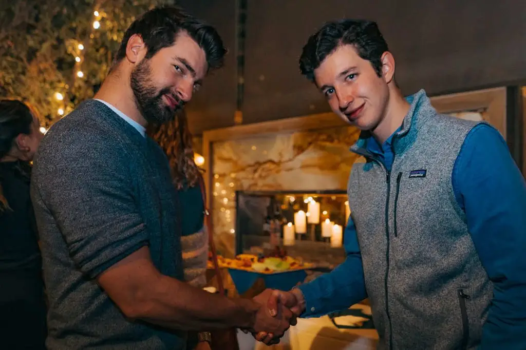 Two men sitting at a high-top table, talking and laughing over drinks and snacks at a local venue.